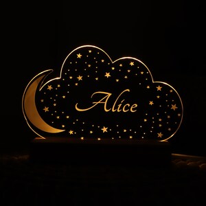 Puede incluir: Una luz nocturna de madera con forma de nube con una luna creciente y estrellas. El nombre "Alice" est&aacute; escrito en una fuente decorativa dentro de la nube.