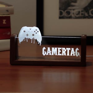 Könnte beinhalten: Eine LED-Leuchte mit Holzrahmen, die das Wort "GAMERTAG" in Weiß anzeigt. Ein weißes Controller-Design mit Tropfeffekt befindet sich über dem Text. Die Leuchte ist eingesteckt und steht auf einer Holzoberfläche.