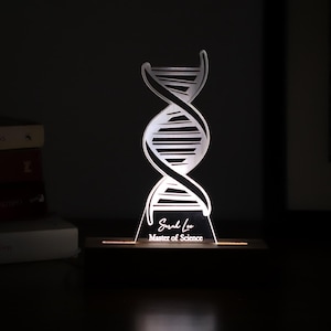 Könnte beinhalten: Eine leuchtende DNA-Helix-Skulptur mit der Inschrift "Sarah Lee Master of Science". Die beleuchtete Doppelhelix steht auf einem Holzsockel und bildet einen Kontrast zum dunklen Hintergrund. Ein einzigartiges Geschenk für einen Absolventen.