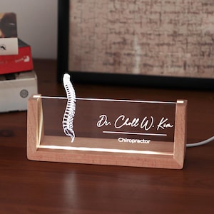 Puede incluir: Un letrero de escritorio rectangular de acrílico transparente con una base de madera iluminada. El letrero presenta un gráfico blanco grabado de una columna vertebral humana a la izquierda y el texto "Dr. Choll W. Kim" sobre "Chiropractor".