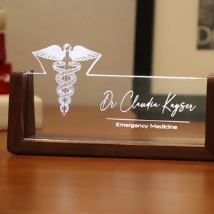 May include: A wooden desk nameplate with a clear acrylic insert. The insert features a white medical symbol and the text "Dr. Claudia Kayser, Emergency Medicine".