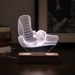 May include: A white 3D printed lamp shaped like a reclining figure with a wooden base. The lamp is lit and casts a warm glow.
