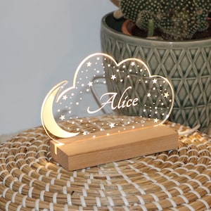Puede incluir: Una luz nocturna de acr&iacute;lico blanco con forma de nube con estrellas y una luna creciente. El nombre "Alice" est&aacute; grabado en la nube. La luz se coloca sobre una base de madera.