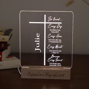 May include: A white acrylic light-up sign with a cross and the text "So loved... Every Day God thinks of you. Psalm 68:19 Every Hour God looks after you. 2 Thessalonians 3:3 Every Minute God cares for you. 1 Peter 5:7 Because Every Second He loves you. Jeremiah 31:3" and the name "Julie". The sign is on a wooden base with the text "Baptized on May 13th 2024".