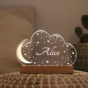 Puede incluir: Una luz nocturna de acr&iacute;lico blanco con forma de nube con una luna creciente y estrellas. El nombre "Alice" est&aacute; escrito en cursiva en la nube. La luz nocturna est&aacute; sobre una base de madera.