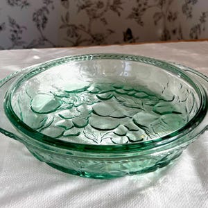 May include: A clear, teal-coloured glass serving dish with a decorative leaf pattern. The dish has a round shape with a slightly raised rim and handles. The glass has a textured appearance, and the dish is on a white surface.