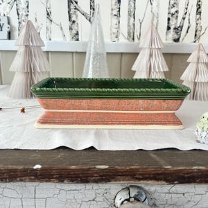 May include: A rectangular ceramic planter with a green rim and a coral-colored body. The planter sits on a white surface with decorative paper Christmas trees and a small bird figurine. A sign that says "THIS IS OUR HAPPY PLACE" is in the background.