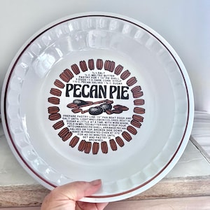 May include: A white ceramic pie dish with a fluted rim and a brown border. The centre features a printed pecan pie recipe, the text "PECAN PIE," and a circular graphic of brown pecan halves. The instructions include ingredients and baking steps.