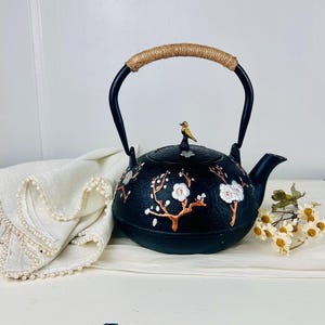 Peut inclure: Une théière en fonte noire avec une surface texturée ornée de branches cuivrées en relief et de fleurs blanches. Le couvercle est surmonté d'une petite figurine d'oiseau doré, et la anse est recouverte d'une cordelette tressée beige clair.