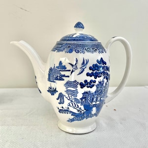 Puede incluir: Tetera de cerámica blanca con un diseño azul de patrón Willow. La tetera tiene un pico curvo, un cuerpo redondeado y un asa. La tapa tiene un pequeño pomo. El diseño azul y blanco representa un paisaje con pájaros y edificios.