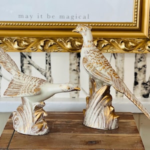 Può includere: Due statuine di uccelli in ceramica bianca con dettagli dorati. Gli uccelli sono appollaiati su una base con un motivo a foglie. Il testo "may it be magical" è visibile sullo sfondo.