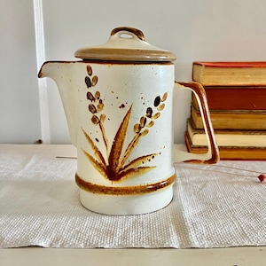 May include: A vintage ceramic coffee pot with a lid. The pot is white with brown floral designs and accents. It has a handle and a spout. The lid has a small handle. The pot is sitting on a white surface.