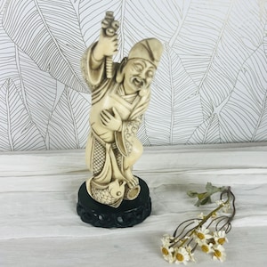 Peut inclure: Une figurine blanc cassé représentant un personnage debout portant une coiffe et des vêtements traditionnels, tenant un bâton d'une main et posant l'autre sur la poitrine. La figurine repose sur un socle noir rond près d'un poisson et de fleurs.