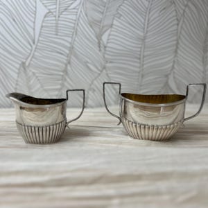 May include: A silver creamer and sugar bowl set. The creamer has a spout and a handle, while the sugar bowl has two handles. Both pieces have a ribbed design on the lower half and a shiny finish.