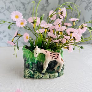 May include: A small, green ceramic planter with a giraffe figurine on the side, filled with pink daisy-like flowers. The giraffe is white with brown spots. The flowers have yellow centers and green stems.