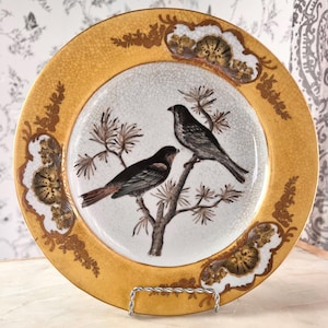 Puede incluir: Plato decorativo con borde dorado y una imagen central de dos pájaros posados en una rama. El plato tiene un fondo blanco craquelado y detalles florales dorados.