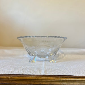 May include: A clear, footed glass bowl with a scalloped rim and floral etching. The bowl has three small feet and a delicate design, perfect for serving small portions or as a decorative piece. The bowl is likely vintage.