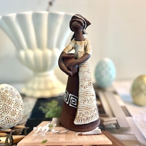 Könnte beinhalten: Keramikfigur einer Frau, die einen Topf hält, mit braunem Rock und weißem Oberteil. Die Figur hat eine Kopfbedeckung und steht vor einer weißen Vase und dekorativen Eiern. Die Eier sind mit floralen Mustern und Goldfarbe verziert.