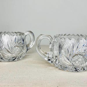 Peut inclure: Deux pièces de service en verre taillé transparent. L'une est une crémière avec un bec verseur, l'autre est un sucrier avec des anses. Les deux présentent un motif en étoile et des bords festonnés. Les pièces sont probablement vintage.