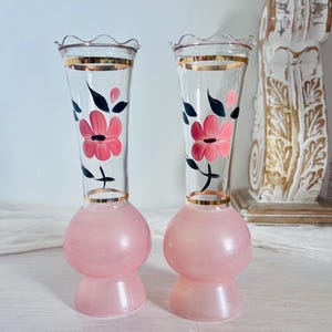 Puede incluir: Un par de jarrones de cristal transparente con bordes ondulados y bandas doradas. La base de cada jarrón es de cristal rosa opaco, mientras que la parte superior presenta flores rosas pintadas a mano con hojas verde oscuro.