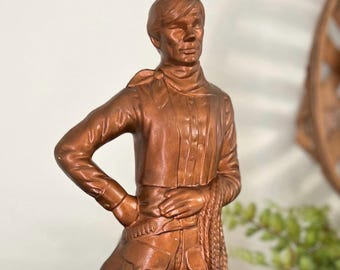 May include: A brown vintage cowboy figurine. The statue depicts a man in a button-down shirt, neckerchief, and cowboy trousers with pockets. He is holding a rope and has one hand on his hip. The figurine is a decorative item.