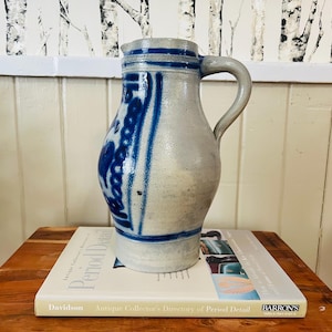 May include: A blue and white ceramic jug with a traditional German design. The jug has a wide base and a narrow neck with a handle.