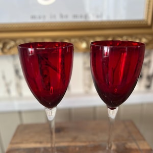 May include: Two ruby red wine glasses with long, clear stems. The glasses have a classic tulip shape and are made of a glossy, transparent material. The background includes a gold framed picture.