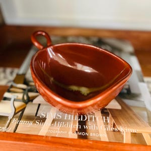 May include: A reddish-brown ceramic gravy boat with a handle. The boat has a smooth, glossy finish and a slightly textured rim. The boat is sitting on a wooden tray with text that reads "Emma Sims-Hilditch with Giles Kime".