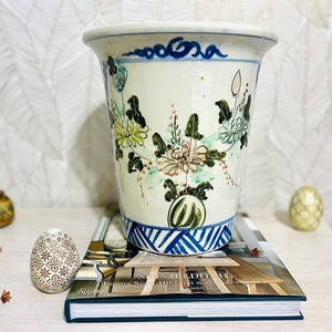May include: A white ceramic planter with hand-painted floral designs in green, yellow, and pink. The planter has a blue and white geometric pattern at the base and a blue wave design at the rim. It is sitting on a book.