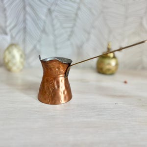 May include: A copper cezve, a traditional Turkish coffee pot, with a long handle. The pot has a hammered texture and a decorative pattern. The cezve is on a light-colored surface, with a blurred background featuring a decorative egg and a small golden object.