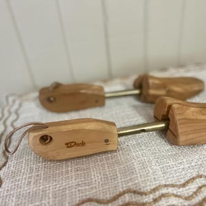 May include: Wooden shoe trees with brass-colored metal rods. The shoe trees are light brown and have a leather loop. The word "Dack" is printed on one of the shoe trees. The shoe trees are on a white and beige patterned surface.
