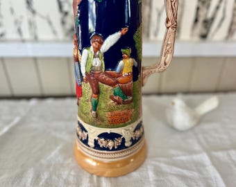 May include: A tall, ceramic beer stein with a detailed relief scene of people dancing in traditional clothing. The stein has a dark blue background, a tan base, and a decorative handle. The text "Tanz auf der Alm" is visible.