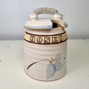 May include: A cylindrical ceramic honey pot with a cream-colored glaze, featuring a lid with a handle and a cutout for a dipper. The word HONEY is embossed in dark brown letters on a band, and a painted blue leaf design with yellow berries is visible.