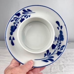 Peut inclure: Un ensemble tasse à thé et soucoupe en céramique blanche. L'ensemble présente un motif floral bleu sur les bords. La tasse à thé est au centre de la soucoupe. L'ensemble est tenu par une main.