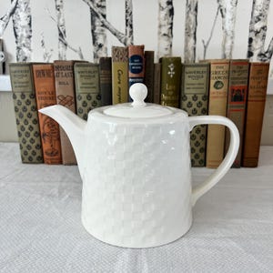 May include: A white ceramic teapot with a textured basketweave design. It features a curved spout, handle, and a lid with a small knob. The teapot sits on a white tablecloth, with a row of vintage books in the background.
