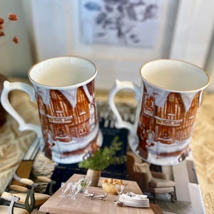 May include: Two white ceramic mugs with gold rims and ornate handles. The mugs feature a detailed illustration of a building with the text "The White Horse" on the front, with a snowy winter scene.