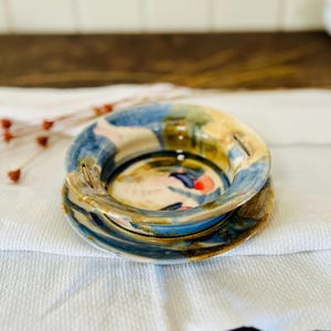 May include: Handmade ceramic bowl and saucer set. The bowl features a colorful abstract design with blue, yellow, and red accents. The set is placed on a white cloth, showcasing the unique artistry of the pottery.