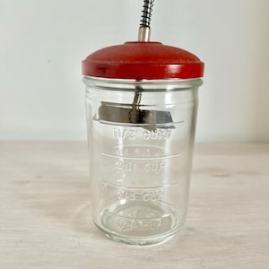 以下が含まれることがあります： 1/3 cup、one cup、2/3 cupの目盛りが刻印された透明なガラス瓶。赤い金属製の蓋が付いており、中央にはバネ式のプランジャー機構と、材料を混ぜ合わせるための内部金属製ミキシングブレードが備わっています。