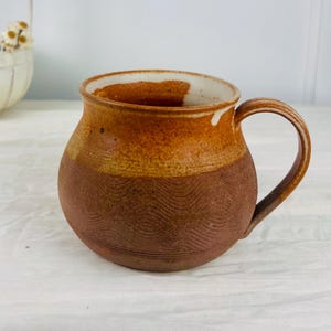 May include: A round ceramic mug featuring a two-tone design with a smooth, glossy amber-coloured glaze on the upper portion and a textured, matte terracotta-coloured base decorated with a repeating wave pattern and a single curved handle.
