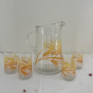 May include: A clear glass pitcher and four matching glasses. The pitcher has a handle and a spout. All the glassware is decorated with a yellow and orange wheat pattern. The glasses are cylindrical and the pitcher is taller.