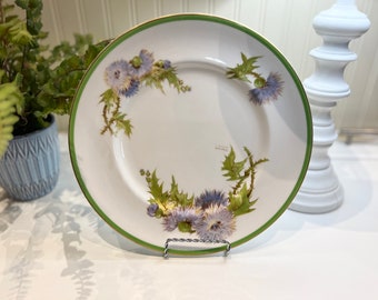 Peut inclure: Assiette en céramique blanche avec un bord vert et une bordure dorée. Elle présente un motif floral de chardons violets et de feuilles vertes. L'assiette est présentée sur un support en fil d'argent, pour la décoration.