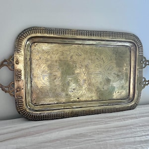 May include: A rectangular brass serving tray featuring an intricate etched floral pattern across the center surface. The tray has a raised, textured border and two ornate, curved handles attached to the sides, showcasing a metallic gold finish.