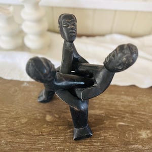 May include: A dark brown wooden sculpture depicting three figures intertwined. The central figure stands upright, supporting two figures that appear to be embracing. The sculpture is set on a wooden surface.