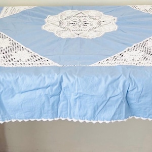 Puede incluir: Un mantel azul claro con detalles de encaje blanco. El mantel presenta un diseño central de encaje floral y detalles triangulares de encaje. Los bordes están acabados con un ribete de encaje festoneado.