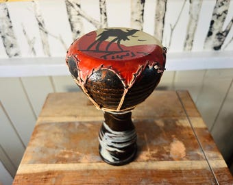 May include: A small, brown, hand-drum with a red and white painted design of a camel in a desert landscape. The drum has a leather top and a wooden base.
