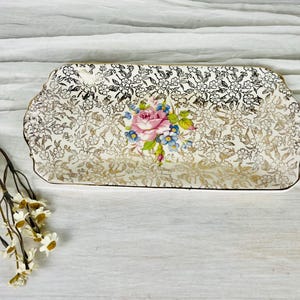 May include: A rectangular ceramic serving tray featuring a white base with an intricate gold floral pattern across the surface and a central bouquet of pink roses and blue forget-me-not flowers with green leaves, accompanied by a sprig of white flowers.