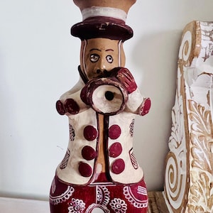 May include: A hand-painted clay figurine of a musician wearing a top hat and a white jacket with maroon buttons, playing a trumpet. The figure features maroon trousers decorated with white floral patterns and geometric lines, standing in an upright position.