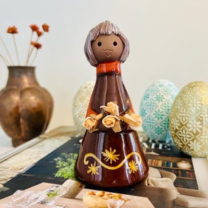 May include: A ceramic figurine of a woman in a brown dress, decorated with yellow flowers. The figurine is set in front of patterned Easter eggs and a copper vase with dried flowers. The figurine is approximately 15 cm tall.
