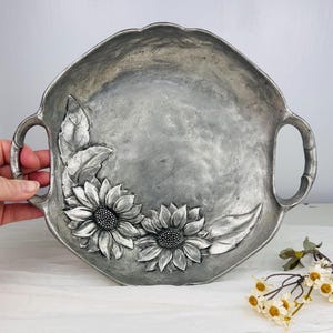 Peut inclure: Un plateau de service en métal argenté avec deux poignées. Le plateau présente des tournesols et des feuilles en relief sur la surface. Le plateau est une pièce décorative pour servir des aliments ou exposer des objets.