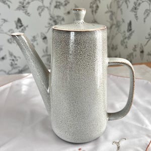 May include: A speckled ceramic coffee pot with a long spout, handle, and lid. The pot is primarily gray with brown accents on the rim, handle, and lid. The background features a floral wallpaper.